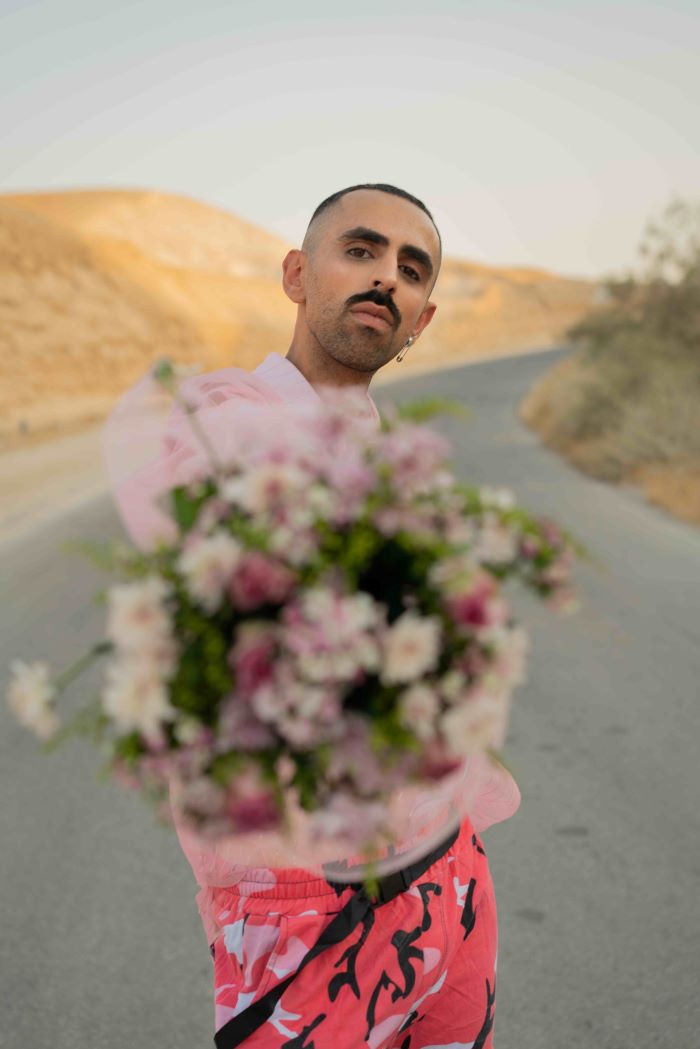 Homme au milieu d'une route tendant un bouquet de fleurs à l'objectif. Paysage derrière lui : montagnes pelées.