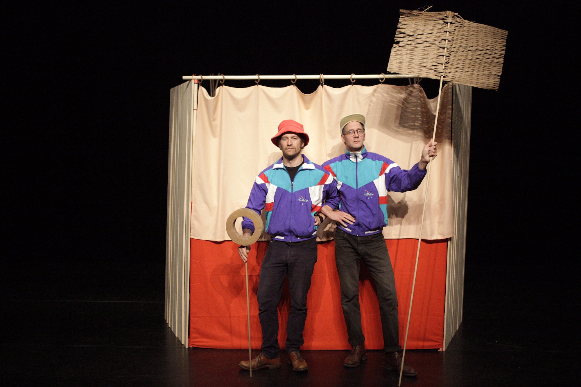 Deux personnages, l'un portant une casquette et l'autre un bob posent dans leur castelet tenant un objet jaune et courbé sur leurs épaules.