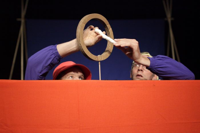 Deux hommes déguisés en enfants jouent avec un bout de plastique en imaginant des histoires improbables.