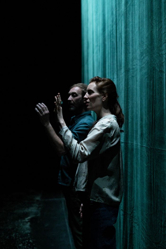 Un homme et un femme sont de profil debout devant un rideau vert. Iels levent toustes les deux la main gauche.