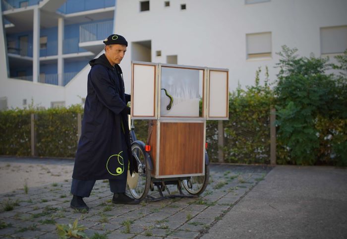 Un homme se trouve derrière un castelet positionné sur un vélo à partir duquel il joue le spectacle dans l'espace public.