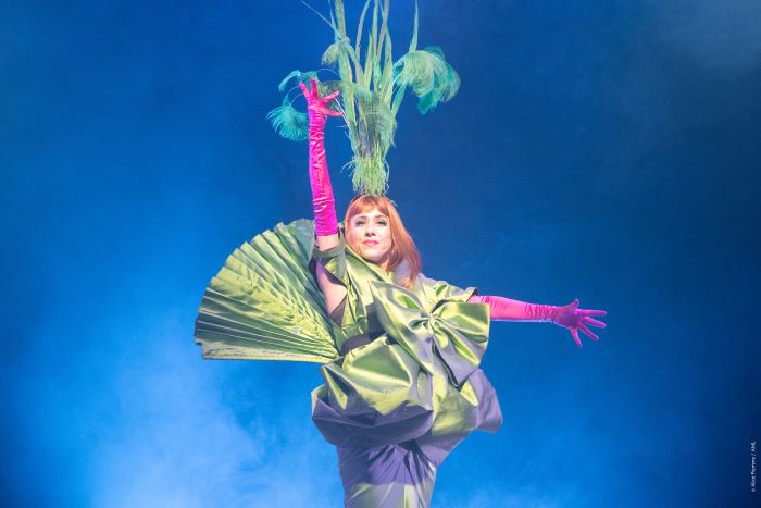 Une danseuse de revue habillée en vert, rose et avec des plumes sur la tête, tend la main droite vers le haut et la gauche sur le côté.