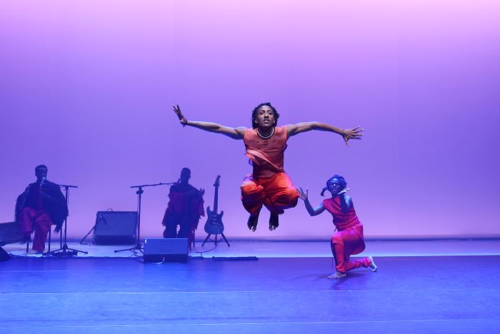 Deux musisciens en fonds de scène et au premier plan deux danseurs en orange dont un en suspension pendant un saut.