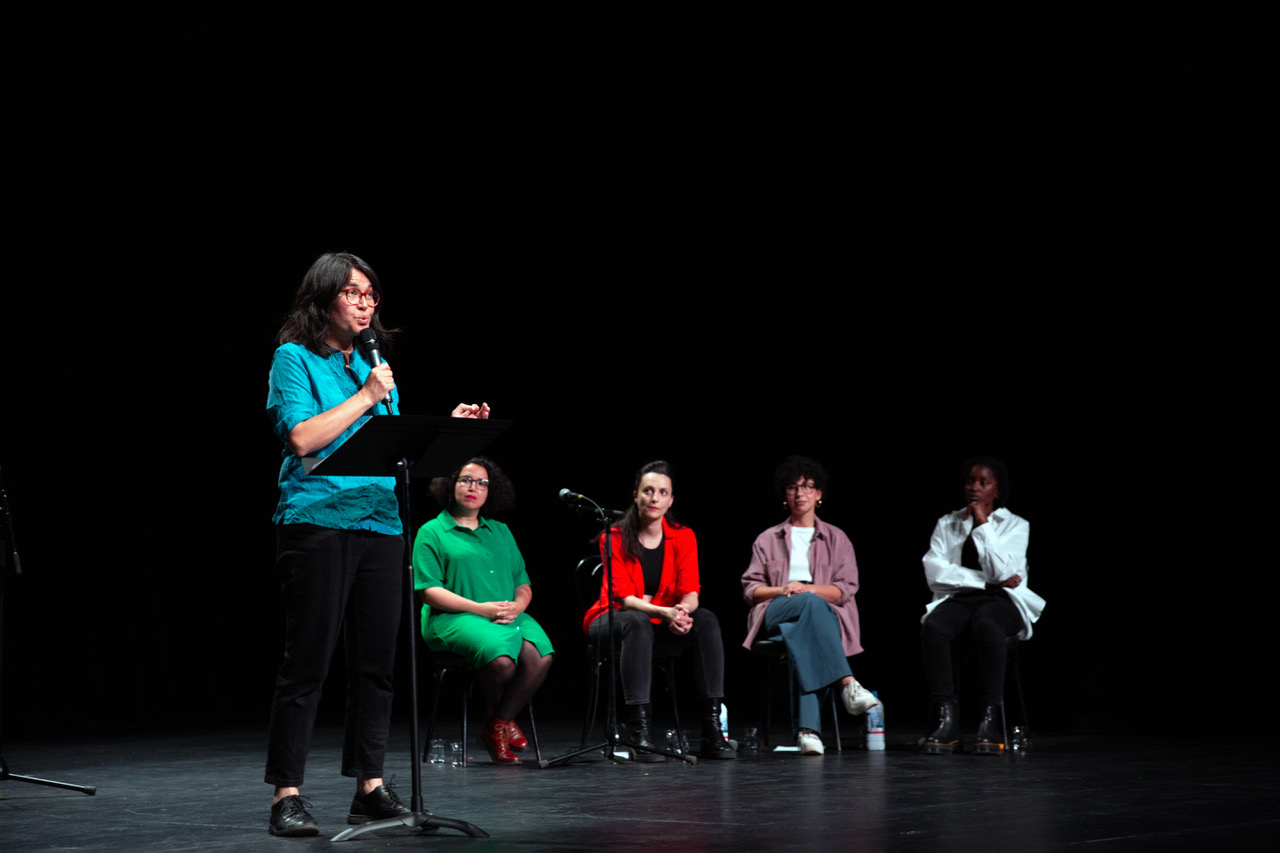 Cinq femmes sur scène. 4 assises, une est au pupitre et parle. 